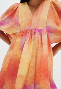 Donna che indossa un vestito fluido con maniche ampie e scollatura a V in un tessuto textured con motivi tie-dye arancioni, rosa e gialli.