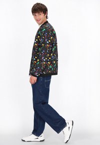 Zwarte bomberjack met een kleurrijk spetterpatroon, gecombineerd met donkerblauwe jeans en witte sneakers. De jas heeft een geribde kraag en manchetten.