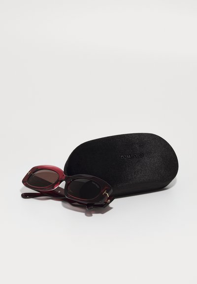 Óculos de sol com uma armação de acetato bordeaux e lentes escuras, repousando ao lado de um estojo preto e texturizado com o logótipo "TOM FORD".