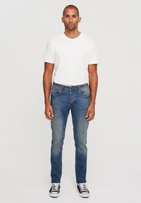 Mėlynos spalvos džinsai iš džinso audinio su siauru kirpimu, šviesiai išblukę ir su siūtomis detalėmis. Derinami su baltu medvilniniu marškinėliu ir juodais žemesniais kedais.