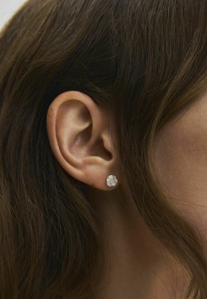 Gros plan sur l’oreille d’une femme aux longs cheveux bruns portant une petite boucle d’oreille argentée ronde en forme de rose.