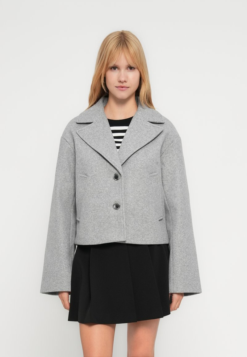 Νεαρή γυναίκα φορώντας μια γκρι μάλλινη κοντή ζακέτα πάνω από μια μαύρη πλισέ φούστα και μαύρο-άσπρο ριγέ τοπ, στέκεται μπροστά σε απλό φόντο.