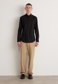 Maglietta nera a maniche lunghe con chiusura a bottoni, abbinata a pantaloni beige chiaro. Modello che indossa scarpe nere, in piedi su uno sfondo neutro.