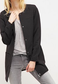 Chaqueta negra con cremallera y cierres a presión, de mangas largas y dos bolsillos frontales. Tela suave con una textura lisa. Diseño de longitud media.