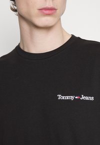 T-shirt en coton noir avec un col rond et un logo "Tommy Jeans" brodé en blanc sur la partie gauche de la poitrine. Texture lisse.