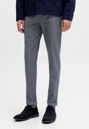 Homme portant un pantalon gris slim, une veste bleu marine, des chaussettes grises et des chaussures en daim noires à lacets, debout devant un fond blanc.