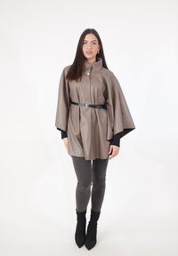 Brauner Cape aus Kunstleder mit hohem Kragen, an der Taille mit einem schwarzen Gürtel geschnürt. Getragen über einem schwarzen Oberteil und grauen Jeans, kombiniert mit schwarzen Stiefeln.