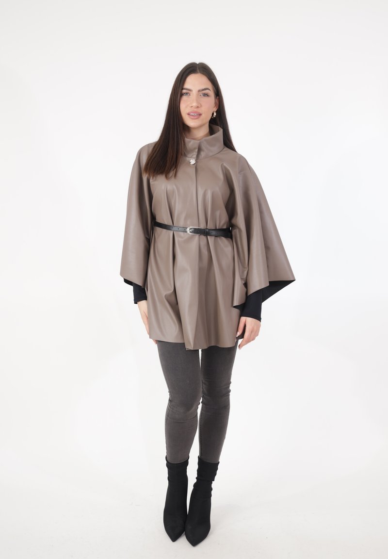 Brauner Cape aus Kunstleder mit hohem Kragen, an der Taille mit einem schwarzen Gürtel geschnürt. Getragen über einem schwarzen Oberteil und grauen Jeans, kombiniert mit schwarzen Stiefeln.