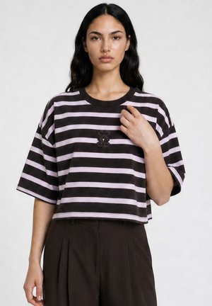 Woman with long dark hair wears a black and light purple striped cropped t-shirt and black pants, standing against a plain background.