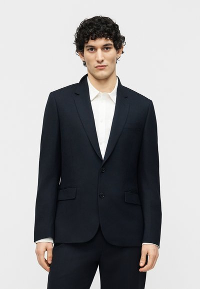 Νέος άνδρας με σγουρά μαλλιά που φοράει μαύρο σακάκι κοστουμιού και λευκό πουκάμισο, στέκεται μπροστά σε απλό ανοιχτό φόντο.