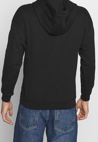 Felpa nera con cappuccio regolabile, materiale misto cotone, polsini e orlo a costine. Indossata con jeans di denim blu, con design delle tasche posteriori.