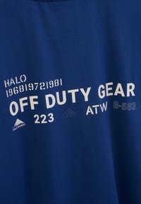 Blå bomullströja med vit text: "HALO 196819721981 OFF DUTY GEAR 223 ATW B-583" med bergslogotyp och prickmönster.