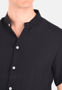 Camicia nera a maniche corte con colletto alto, tessuto testurizzato e bottoni bianchi lungo la pattina anteriore.