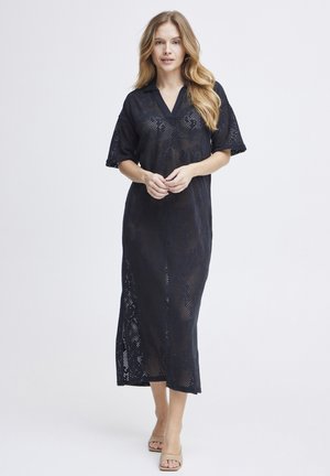 Femme aux longs cheveux portant une robe midi noire perforée à manches courtes et des talons ouverts beige, debout devant un fond blanc uni.