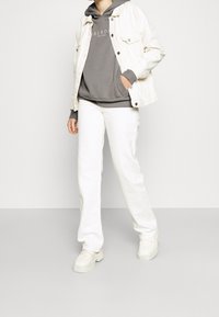 Weiße Jeansjacke, grauer Hoodie mit Logo und weiße schmal geschnittene Hose. Klobige weiße Sneakers runden das Outfit ab. Weiche Texturen im gesamten Look.