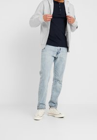 Grauer Kapuzenpullover mit Reißverschluss über einem marineblauen Henley-Shirt, kombiniert mit hellblauen Jeans und weißen Turnschuhen. Verfügt über ein lässiges, lockeres Passformdesign.