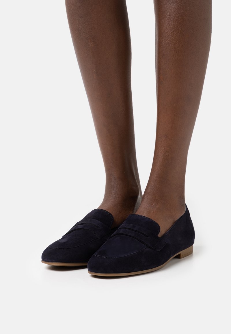 Mocassins en daim bleu marine avec un talon plat, présentant des détails de couture et une texture lisse. Les chaussures sont posées sur un fond clair.
