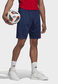 Granatowe szorty sportowe z czerwonymi akcentami po bokach, w połączeniu z białymi sneakersami i skarpetkami. Trzyma wielokolorową piłkę nożną Adidas.