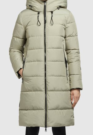 Femme portant un long manteau matelassé beige clair avec capuche, fermeture éclair à l'avant et poches latérales zippées.