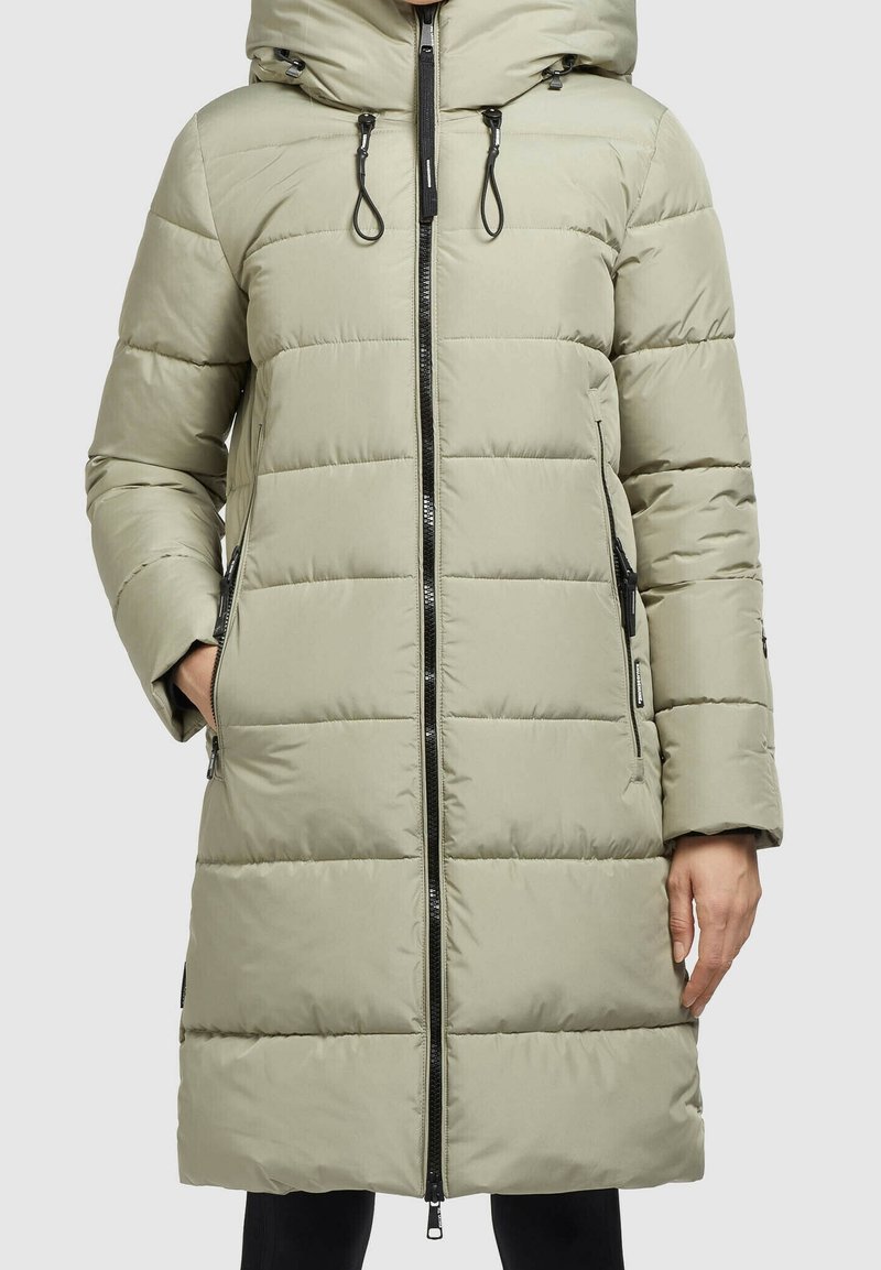 Femme portant un long manteau matelassé beige clair avec capuche, fermeture éclair à l'avant et poches latérales zippées.