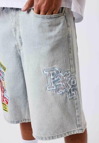 Pantalones cortos de mezclilla clara con parches coloridos y bordados, usados por una persona con la mano derecha en el bolsillo delantero.