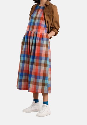 Nainen, joka pukeutuu moniväriseen ruutukuosiseen midimekkoon, ruskeaan takkiin, jossa on käännetyt hihat, sinisiin sukkiin ja valkoisiin korkeavartisiin tennareihin, seisoo käsine taskuissa.