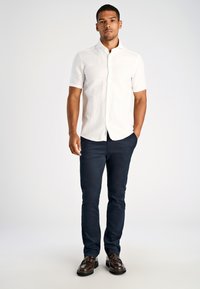 Man staat met één hand in zijn zak, draagt een wit kortpoloshirt met knopen, een donkere broek en bruine tassel loafers.
