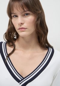 Junge Frau mit welligem braunem Haar, die ein weißes Oberteil mit einem navyblauen und weißen gestreiften V-Ausschnitt trägt, neutrale Gesichtsausdruck, heller Hintergrund.