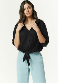 Sort, løstsiddende skjorte med knaplukning og knyttet detalje foran. Har optrillede ærmer og en afslappet krave. Matchet med lyseblå jeans.