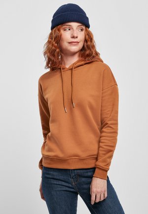Sweat-shirt en coton marron avec un cordon de serrage et des extrémités argentées, associé à un jean bleu foncé. Le modèle porte un bonnet en tricot bleu marine.