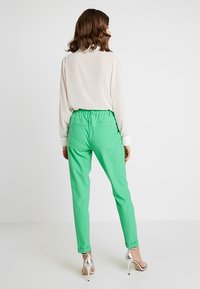 Groene stretchbroeken met een elastische tailleband, gecombineerd met een doorzichtige witte blouse. De outfit bevat metalen hoge hakken.