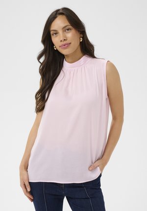 Hosszú sötét hajú nő világos rózsaszín ujjatlan blúzban és sötétkék farmernadrágban, fehér háttér előtt áll.