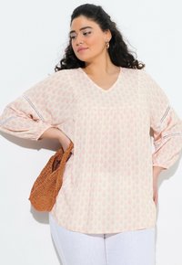 Blusa rosa chiaro con motivo floreale, scollatura a V e maniche larghe con bordino decorativo. Abbinata a una borsa di paglia intrecciata.