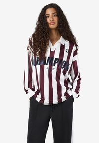 Vrouw met lang krullend haar, gekleed in een bordeauxrode en witte gestreepte lange mouwen shirt met de tekst "CHAMPION" en zwarte broek, met haar handen in haar zakken.