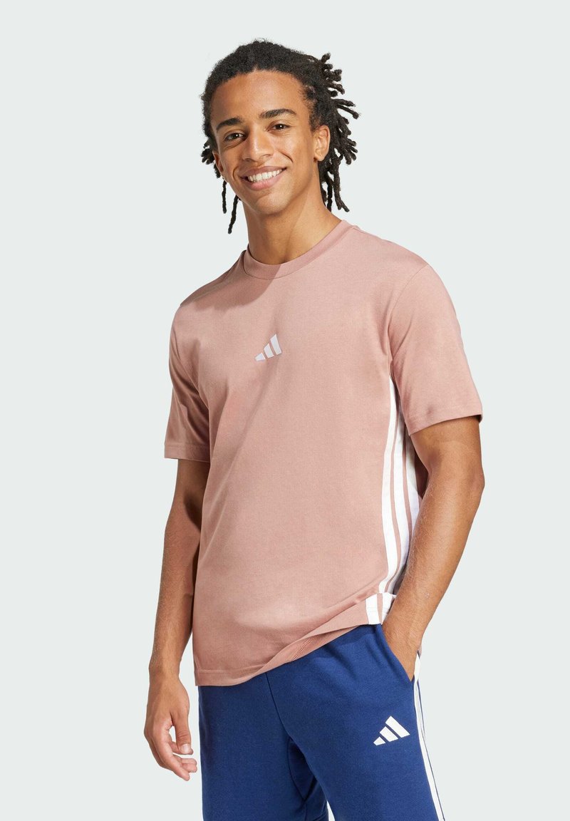 Camiseta de algodón rosa de manga corta con el logo blanco de Adidas en el pecho y rayas blancas en los laterales, combinada con pantalones azul marino.
