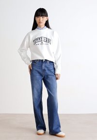 Sudadera blanca con "TOMMY JEANS NEW YORK CITY" en negro, combinada con unos vaqueros de pierna ancha azules y zapatillas blancas con suela color beige.