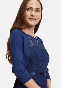 Robe bleu marine avec un corsage en dentelle et des manches trois quarts, présentant une ceinture en satin à la taille et des détails floraux en dentelle intriqués.