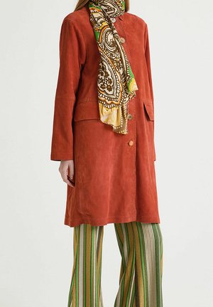 Cappotto in suede color ruggine con chiusura a bottoni, abbinato a una sciarpa a fantasia multicolore e pantaloni a righe verdi a gamba larga.