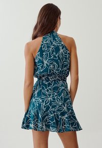 Turkos halterneck-klänning med vitt blommönster, rynkat midja och volangkant, tillverkad av lättviktig tyg. Rygglös design.