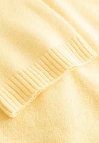 Měkká žlutá pletená látka zobrazující detail žebrovaného lemu, zdůrazňující texturu a vzor stehů teplého rukávu oděvu.