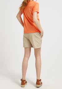 Žena v oranžovém tričku s krátkým rukávem, béžových kraťasech ke kolenům a hnědých sandálech na platformě, stojící s rukama v kapsách, otočená zády.