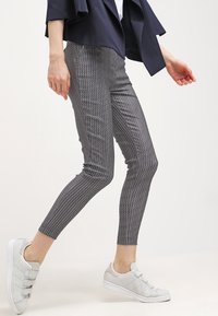 Person som går iförd svart- och vitrutiga slim jeans, marinblå jacka, vit topp och vita sneakers med kardborreband.