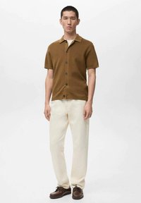 Polo marrone a maniche corte in maglia con colletto, fronte con bottoni, abbinata a pantaloni crema e scarpe marroni, in piedi su uno sfondo neutro.