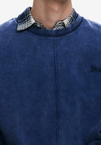 Sweat-shirt bleu marine à la texture douce, doté d'un col avec un motif géométrique en crème et bleu marine, et d'une broderie discrète de la marque.