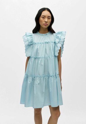 Ung kvinde med langt sort hår iført en lyseblå kjole med lag og bølget flæseskørt mod en ensfarvet hvid baggrund.