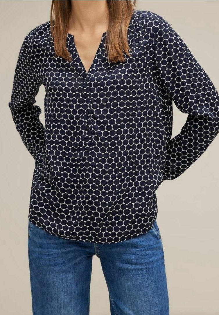 Femme portant un chemisier bleu marine à manches longues avec des pois blancs et un jean bleu en denim, les mains reposant dans son dos.