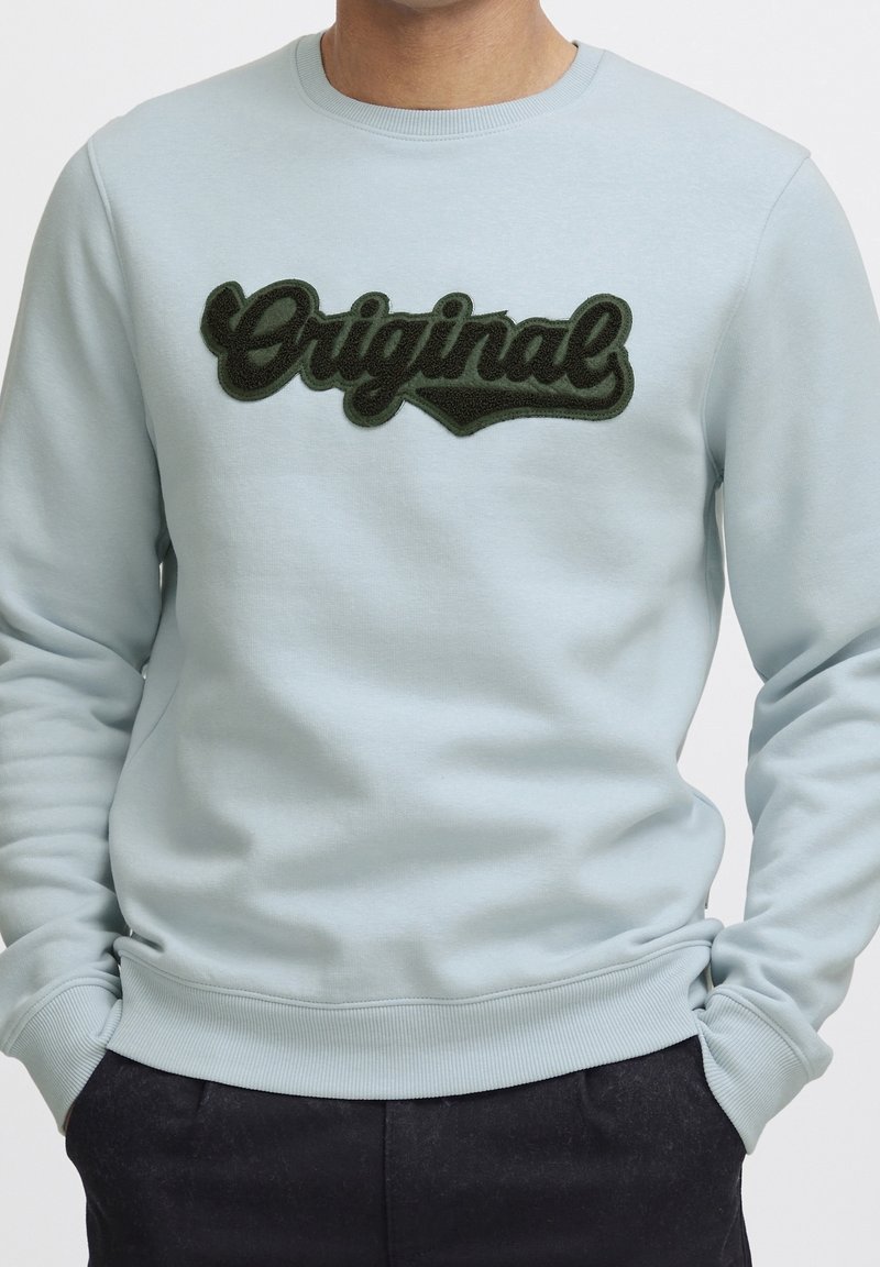 Homme portant un sweat-shirt bleu clair avec un texte "Original" vert foncé sur la poitrine, mains dans les poches, pantalon noir visible.
