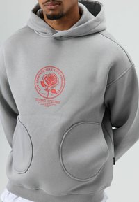 Grijze capuchontrui gemaakt van zachte stof, met een ronde bloemmotief en twee oversized zijzakken met gestikte details.