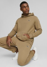 Felpa beige e pantaloni jogger con logo Puma nero, tessuto morbido, tasca frontale e polsini a costine. Il modello indossa sneakers bianche.