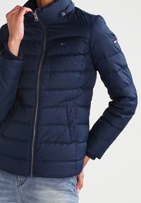Granatowa kurtka puchowa z wysokim kołnierzem, zapięciem na zamek, poziomym przeszyciem i bocznymi kieszeniami. Posiada logo marki na rękawie.